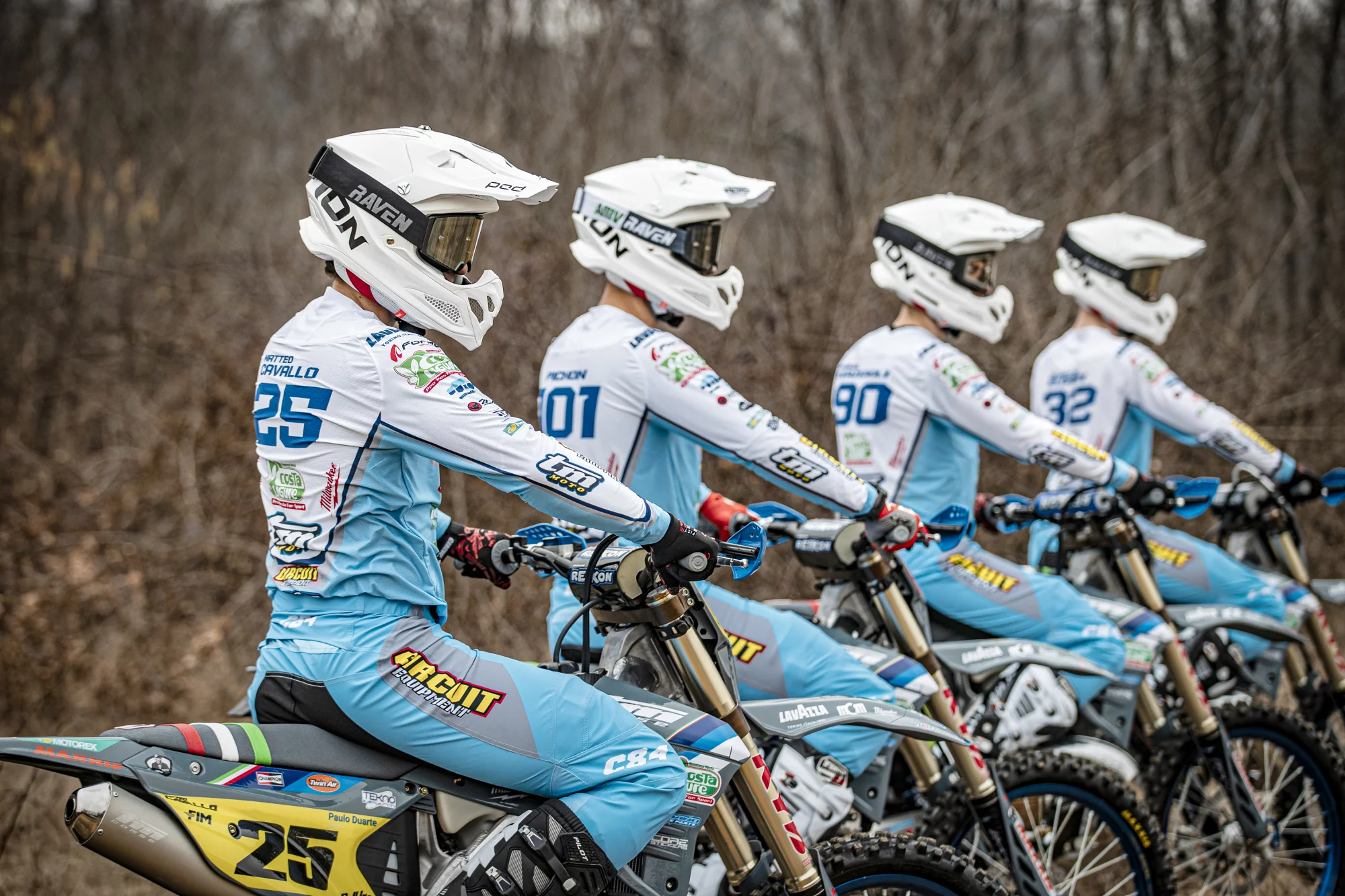 Vier motorcrossers in blauw-witte teamkleding staan zij aan zij op hun motoren in een rij, klaar voor vertrek in een bosrijke omgeving.