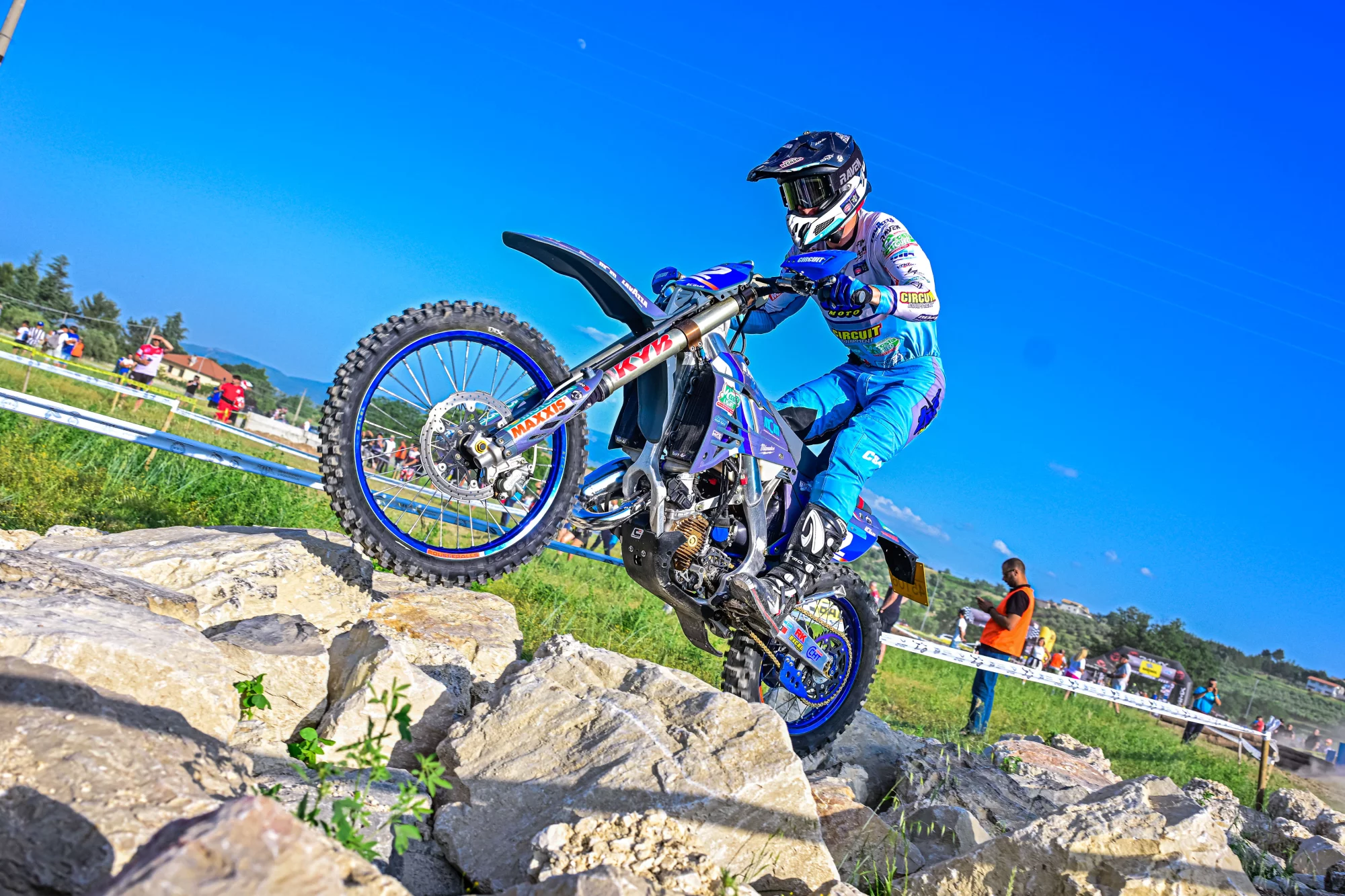 Motorcrosser in blauwe outfit rijdt over een rotsachtig parcours tijdens een wedstrijd op een zonnige dag.