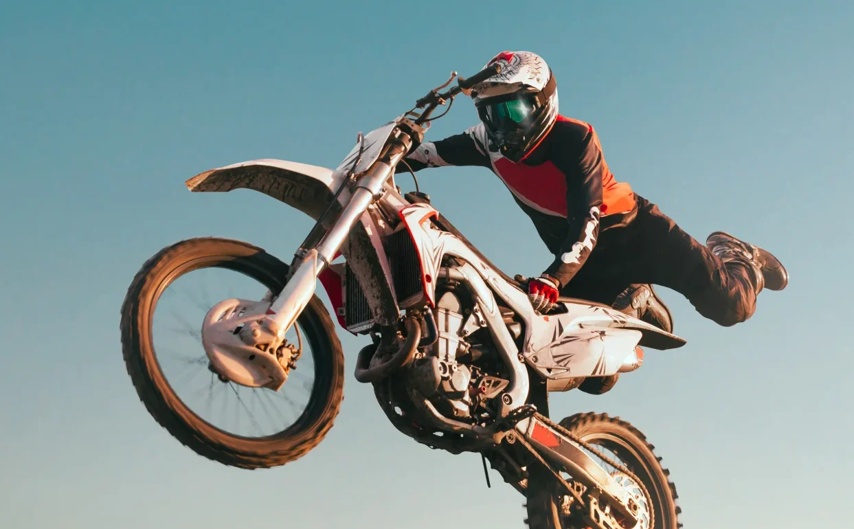 Freestyle motorcrosser voert een trick uit in de lucht met één been van de motor af, tegen een heldere blauwe lucht.
