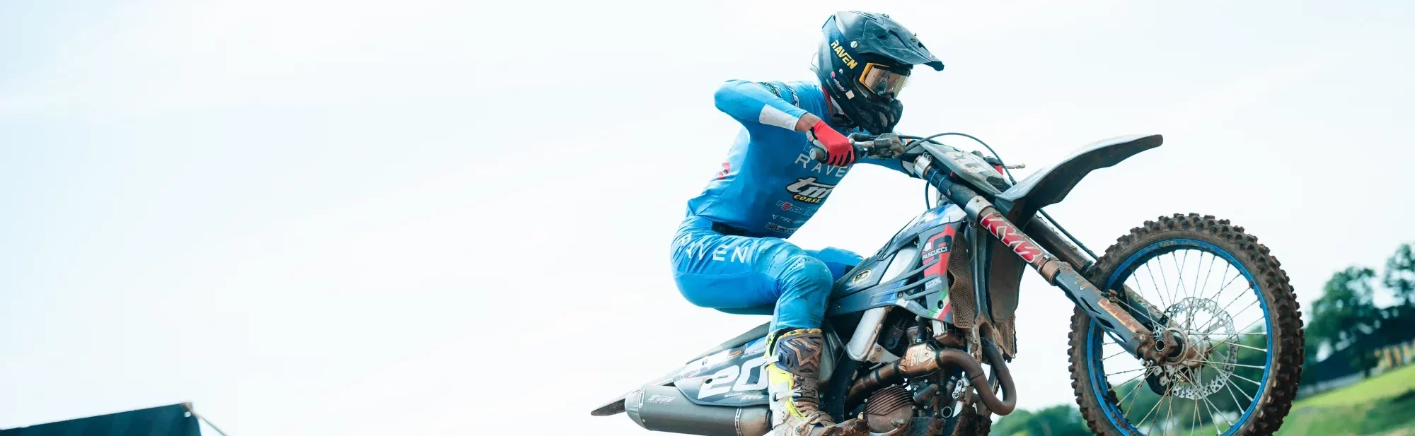 Motorcrosser in blauwe outfit maakt een sprong over een modderige heuvel, met beide wielen los van de grond.
