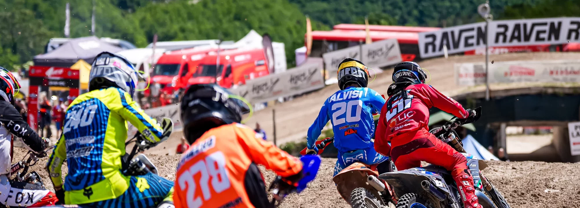 Groep motorcrossers in actie tijdens een race op een zandcircuit, met kleurrijke outfits en achtergrond van tenten en publiek.