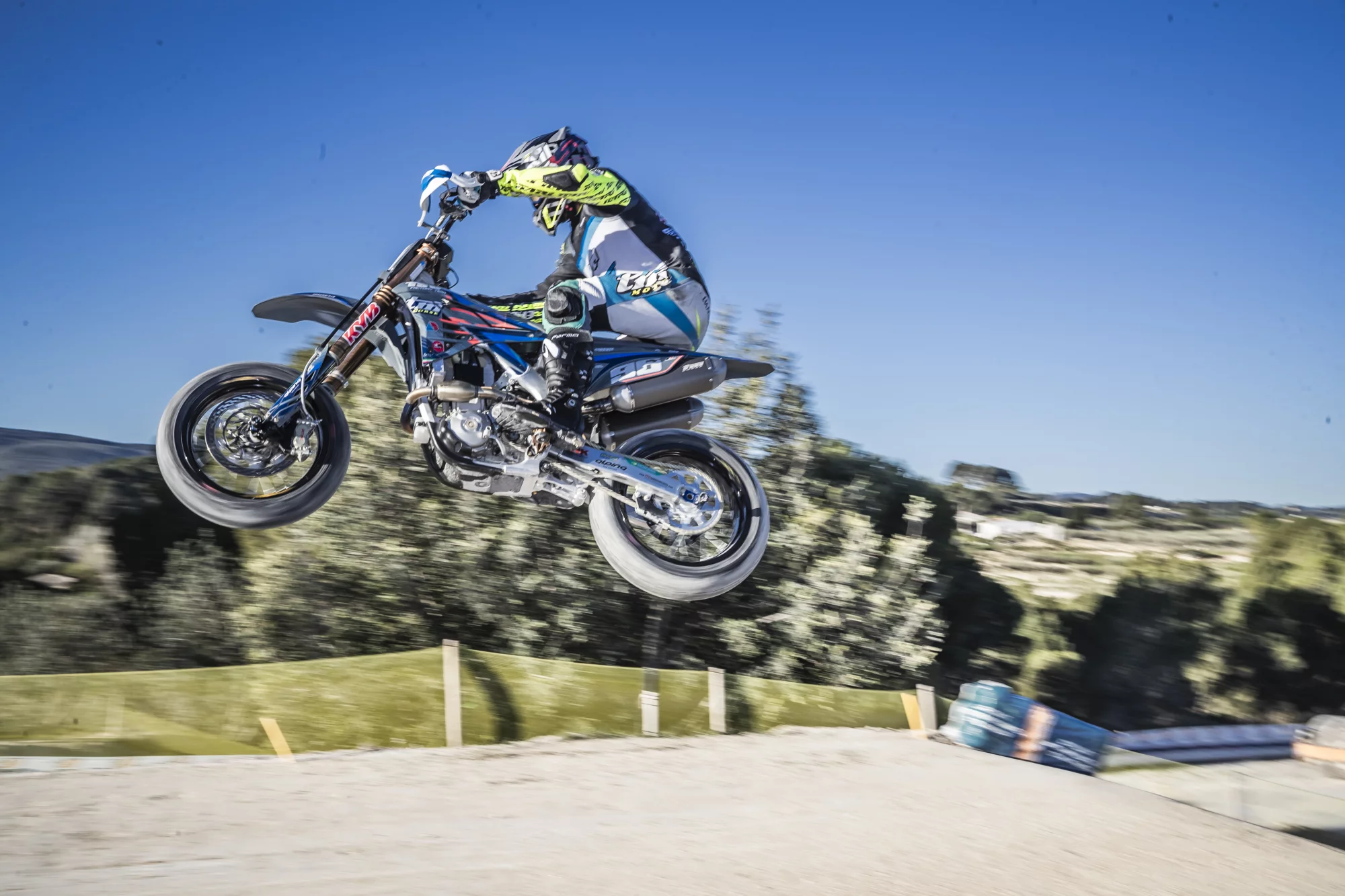 Motorcrosser in kleurrijke outfit springt met hoge snelheid over een heuvel tijdens een zonnige offroadwedstrijd.
