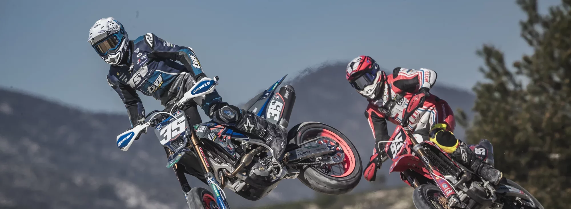 Twee motorcrossers maken een sprong tijdens een race, één in blauwe outfit met nummer 35 en de ander in rood met nummer 95, met bergen op de achtergrond.